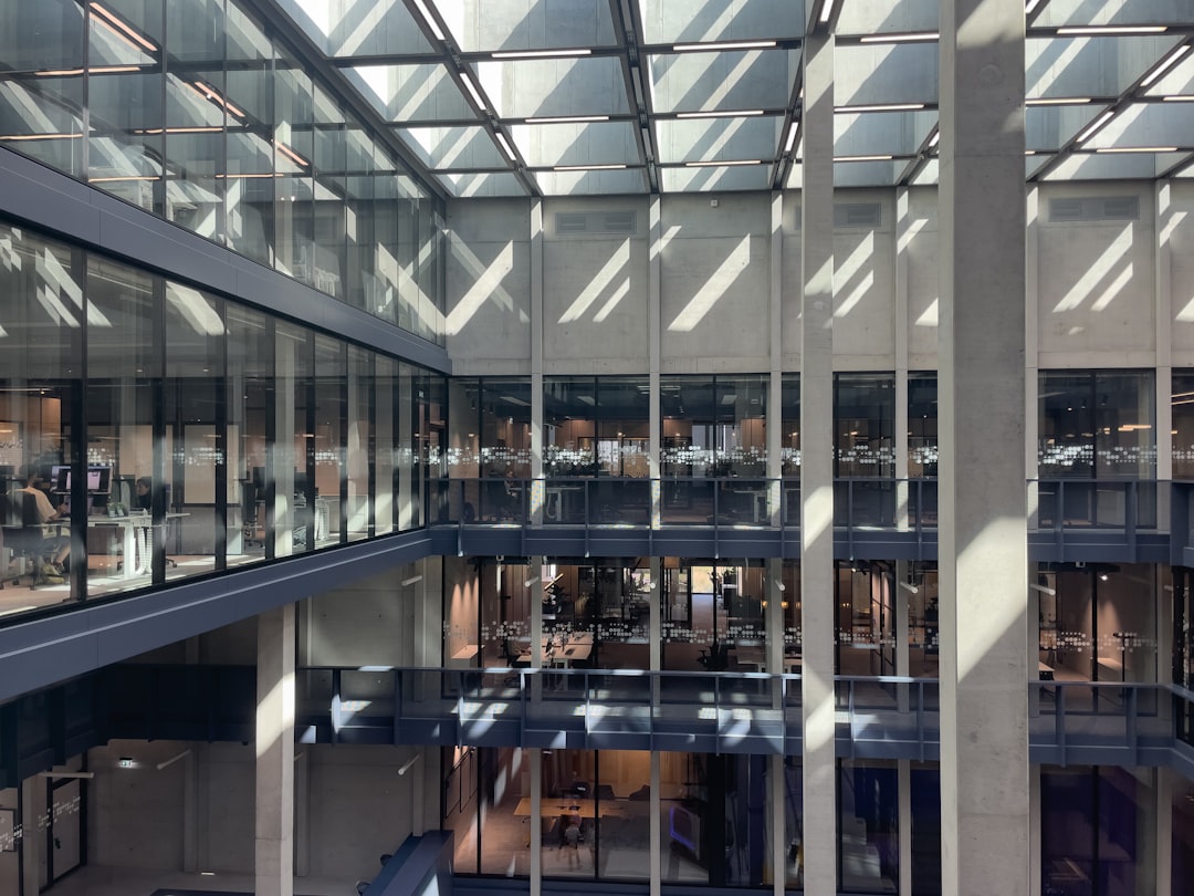 A modern office atrium with a glass ceiling allows natural light to flood the space casting dynamic shadows and reflections on the interior The multi level design features large glass walls and concrete pillars creating an open airy environment Inside workspaces and offices are visible through the glass while a few employees move through the central area emphasizing the buildings functional and contemporary design The architecture blends industrial elements with modern office aesthetics